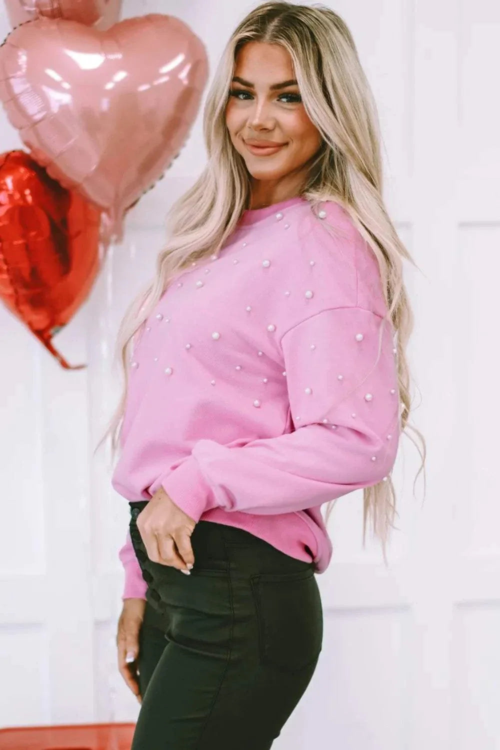 Woman wearing pink pearl round neck dropped shoulder sweatshirt with black pants and heart-shaped balloons in the background