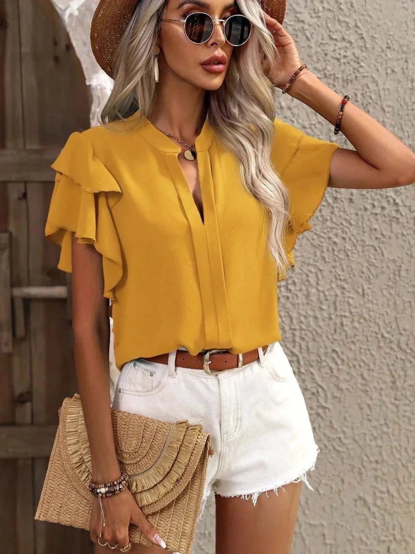 Woman wearing mustard yellow ruffle sleeve notched blouse with white shorts and straw hat holding woven clutch bag