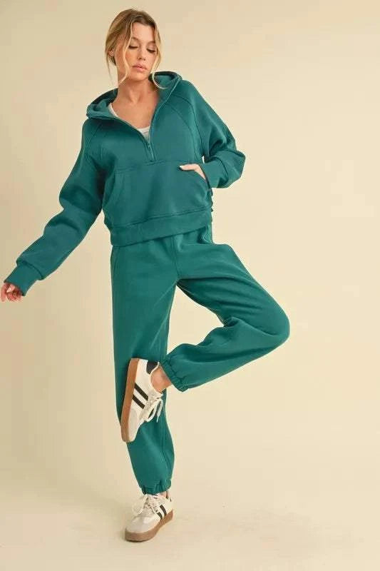 Woman wearing teal Aemi+Co drawstring elastic waist joggers and matching hoodie with white sneakers posing against beige background